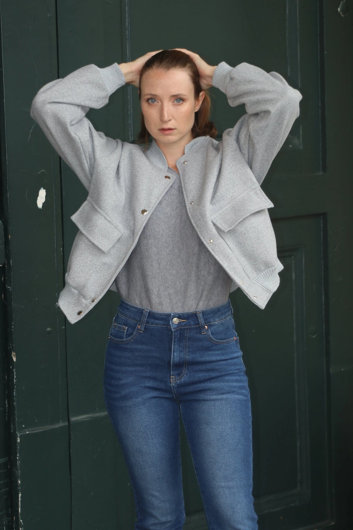 Femme portant un blouson gris clair, un haut gris et un jean bleu devant une porte verte.