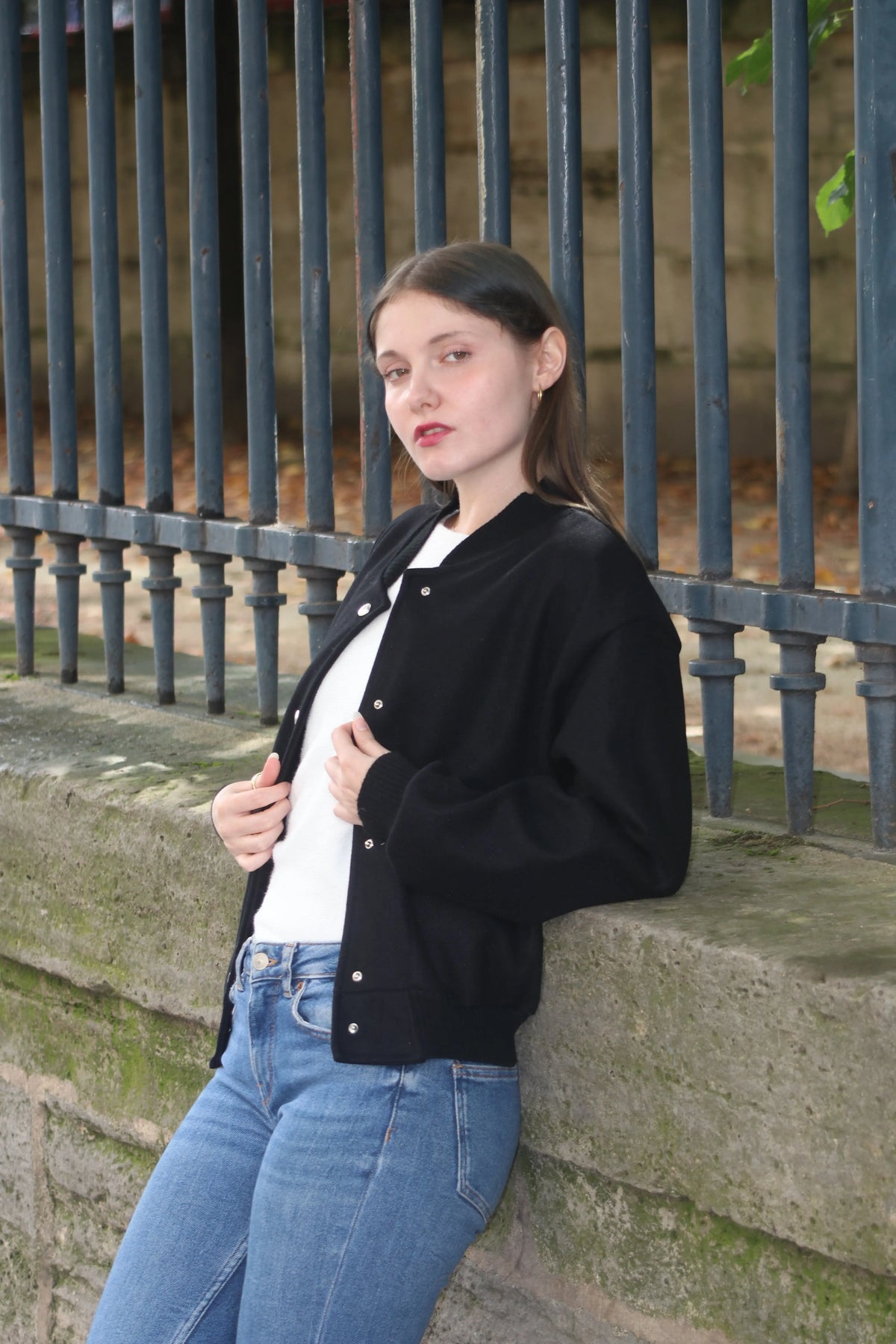 Jeune femme portant une veste noire et un jean bleu, posant devant une clôture en fer.