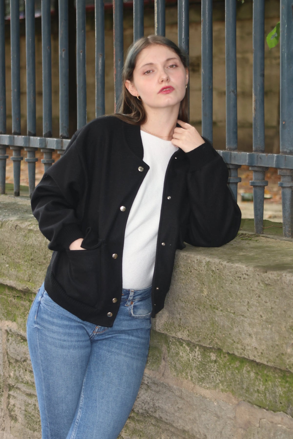 Femme portant un blouson noir et un t-shirt blanc avec un jean bleu, posant devant une clôture.