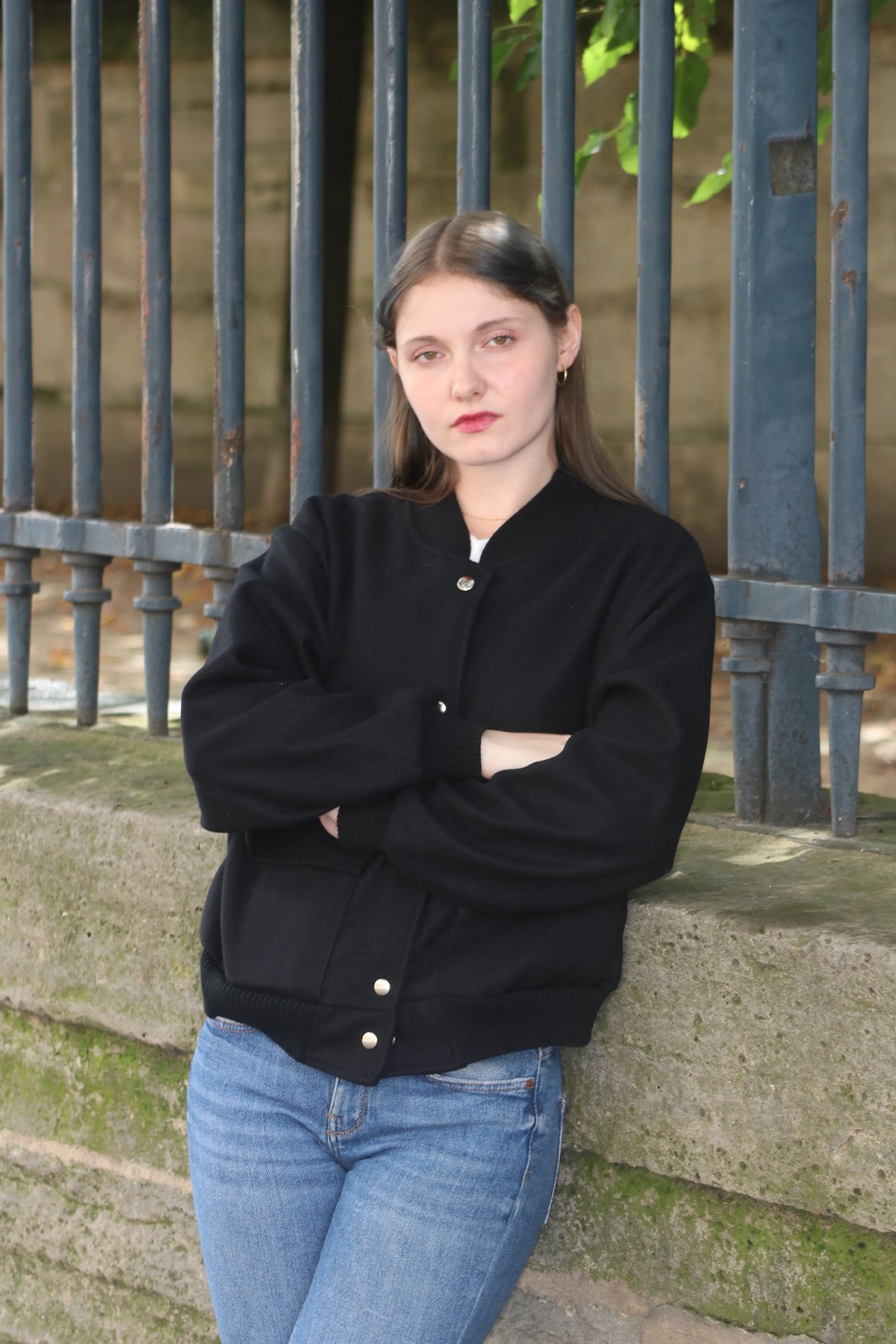 Jeune femme en veste noire et jean bleu posant devant une grille métallique.
