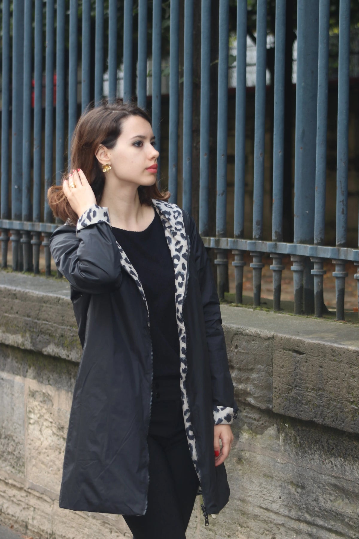 Femme portant une parka noire avec doublure imprimé léopard devant une grille métallique.