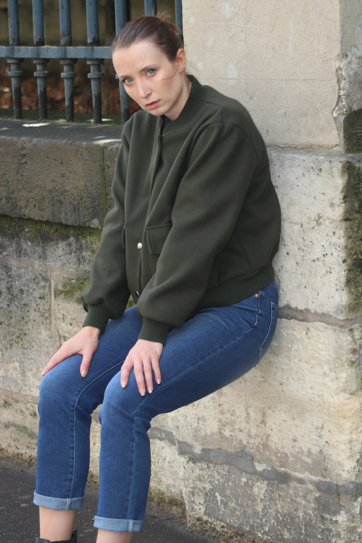 Femme portant une veste bomber vert olive et un jean bleu, assise contre un mur en pierre.