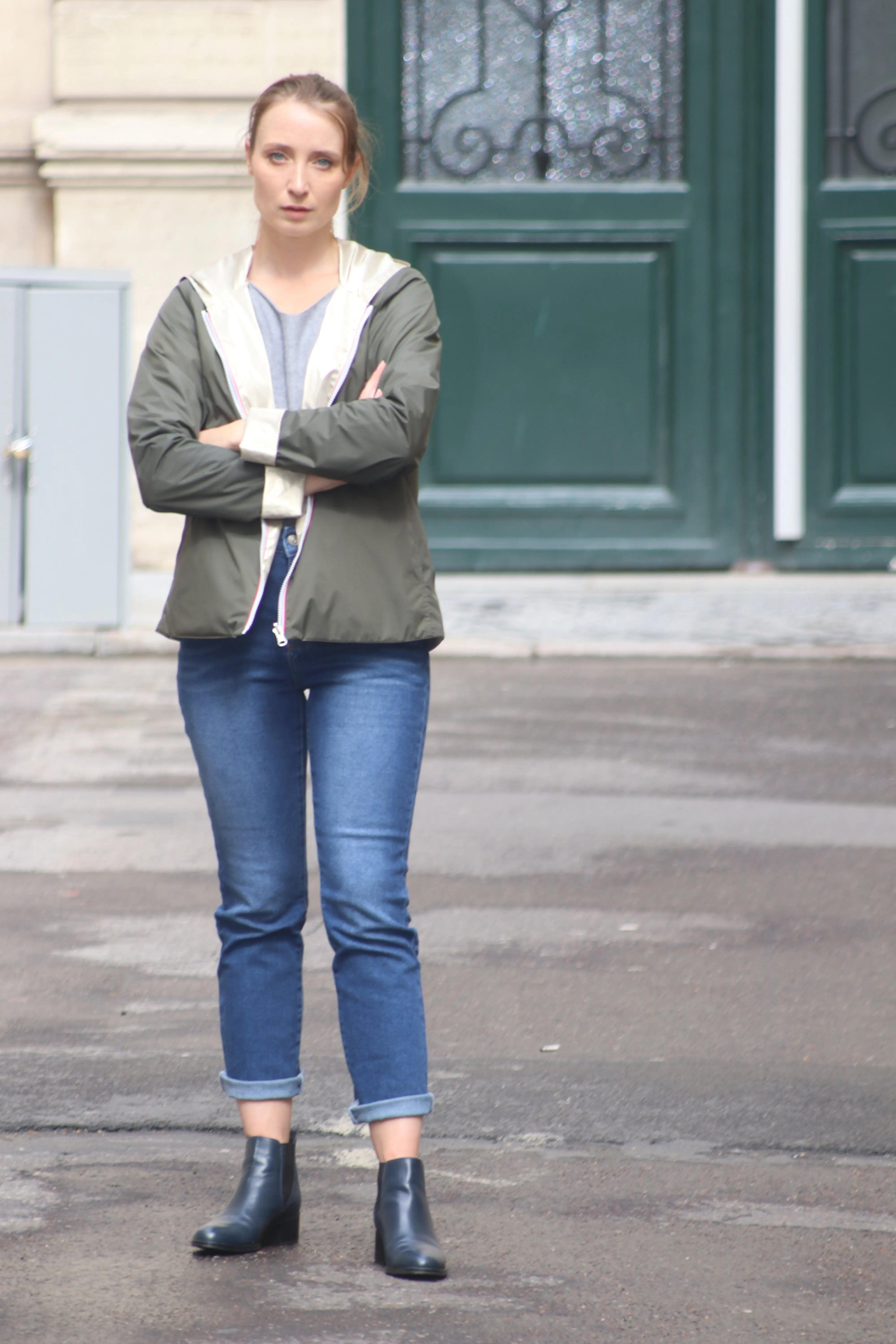 Femme portant une veste olive, un jean bleu et des bottines noires devant une porte verte.