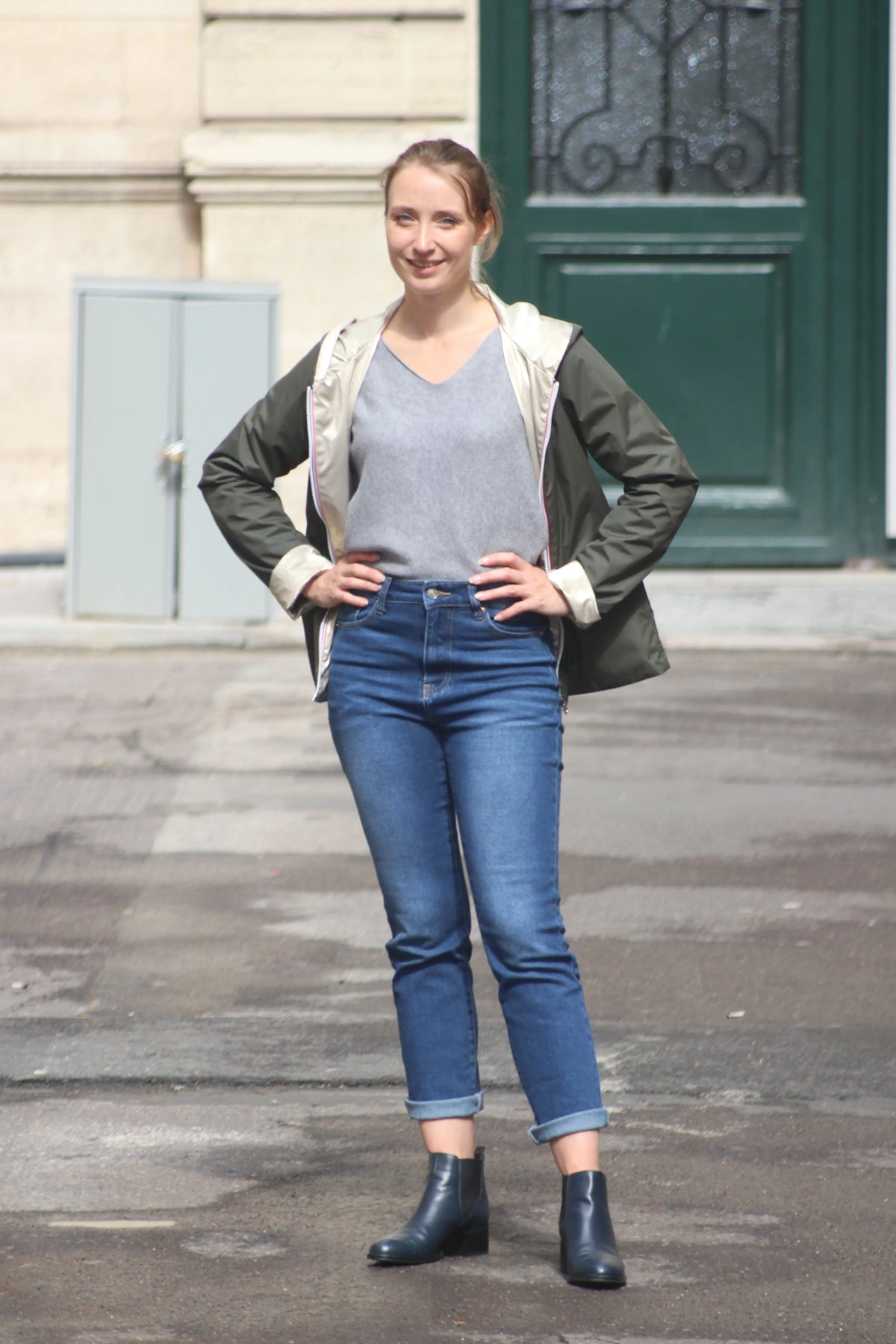 Femme portant un jean bleu, un t-shirt gris et une veste olive sur un fond urbain.
