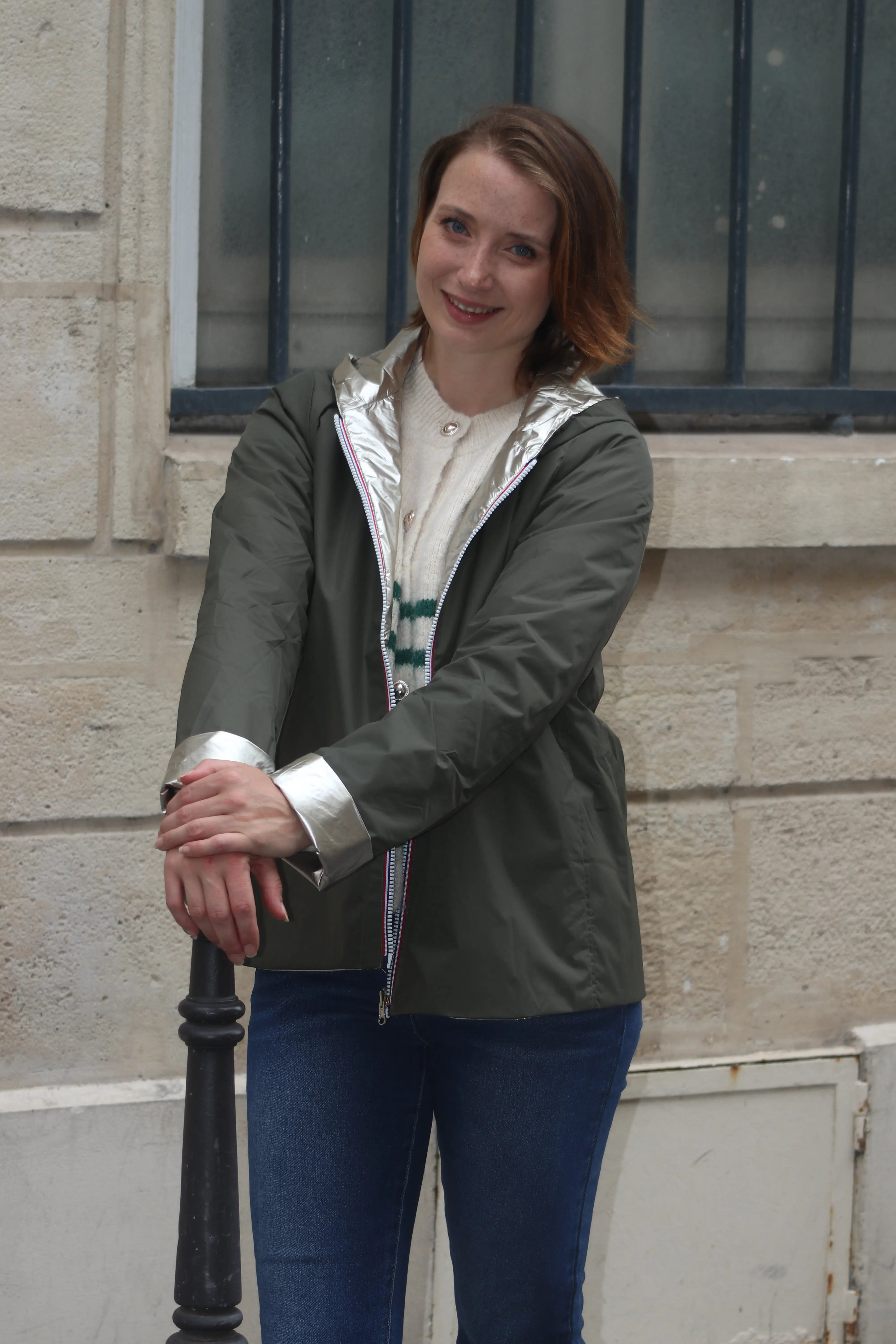 Femme souriante portant une veste olive avec détails argentés et un pull blanc.