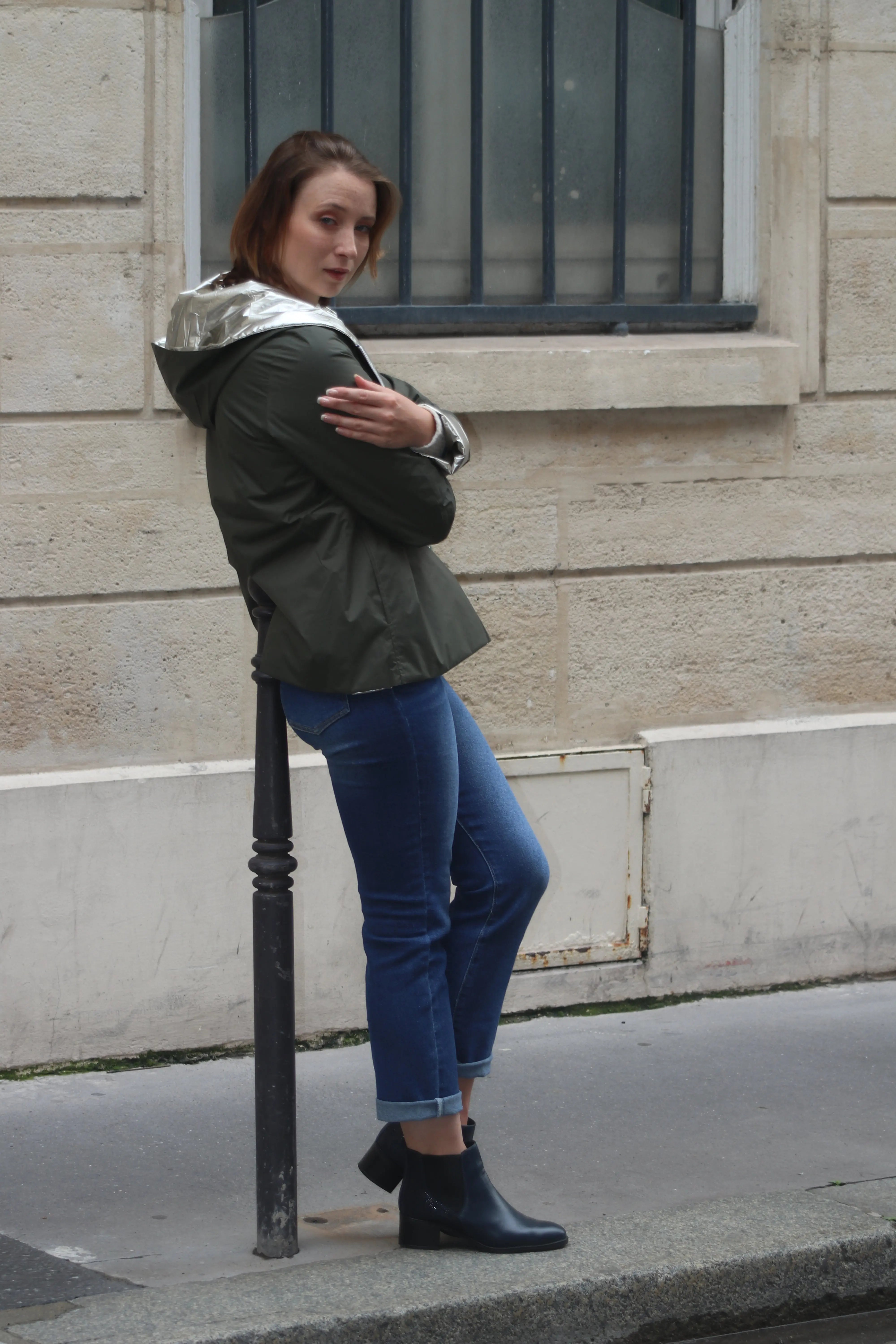Femme portant une parka vert olive avec capuche argentée et jean bleu sur un trottoir parisien.