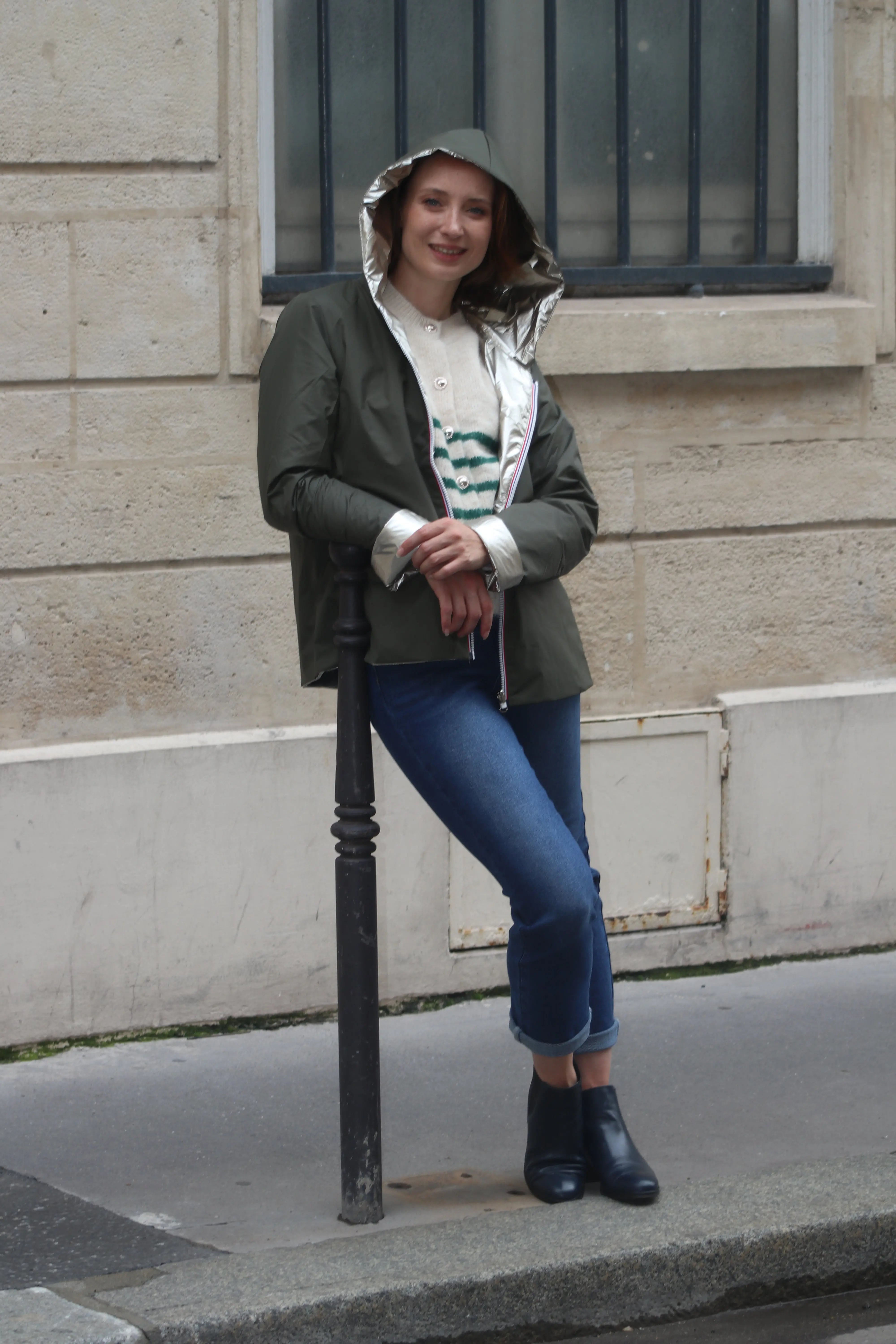 Femme portant un coupe-vent vert olive et un jean bleu devant un mur en pierre.
