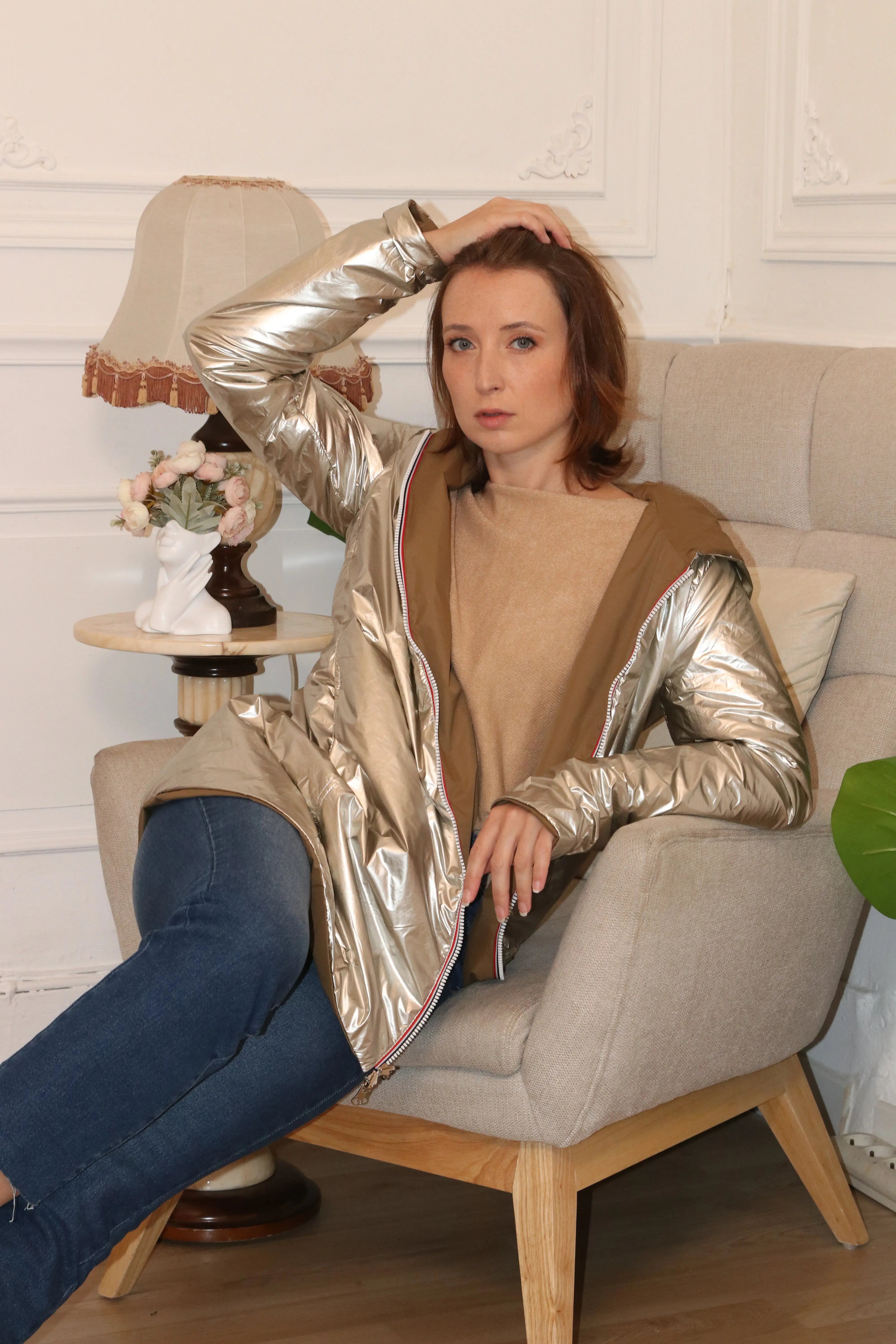 Femme portant une veste légère métallisée dorée et un pull beige assise dans un fauteuil.