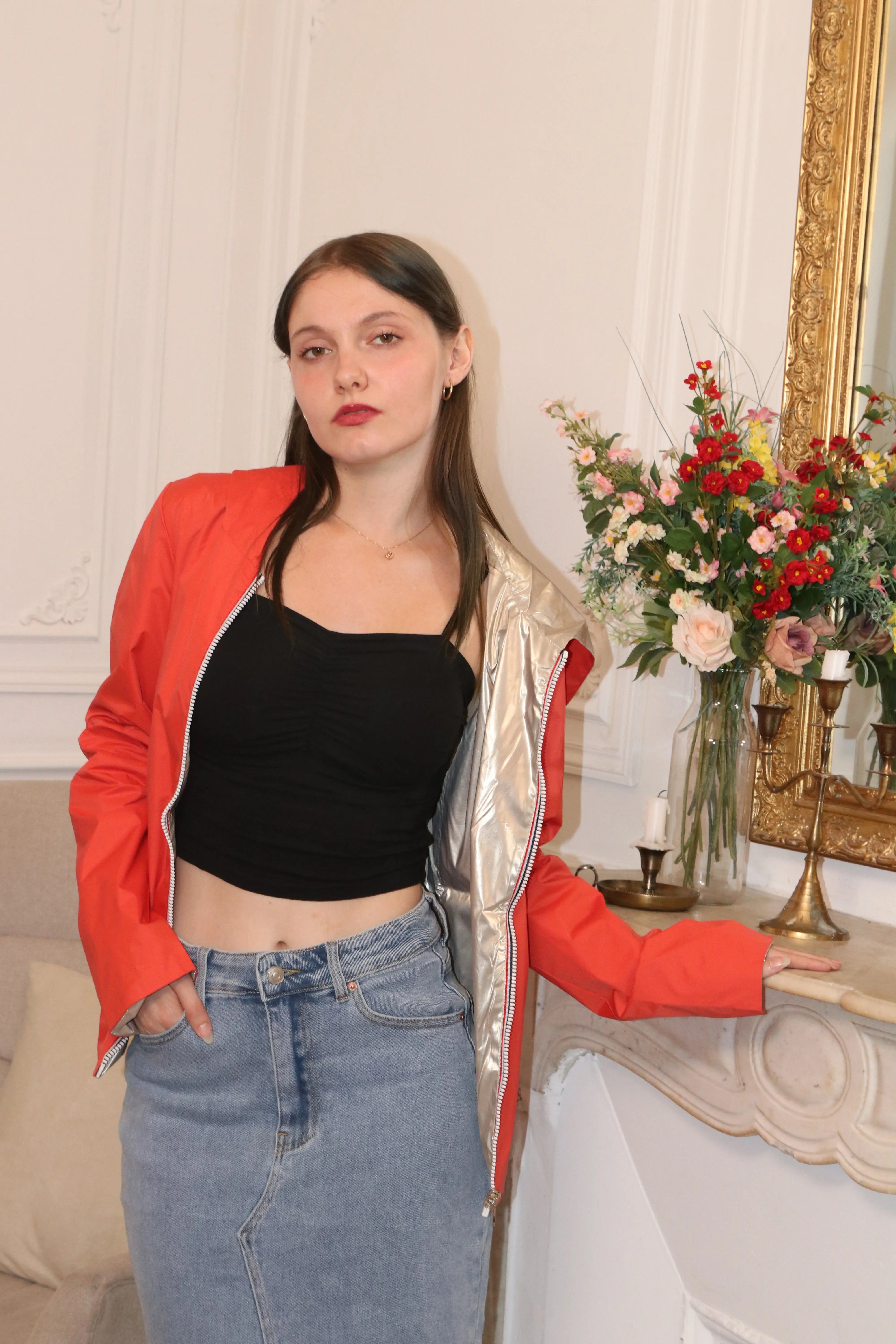 Femme portant une veste rouge, un top noir et une jupe en jean devant des fleurs.