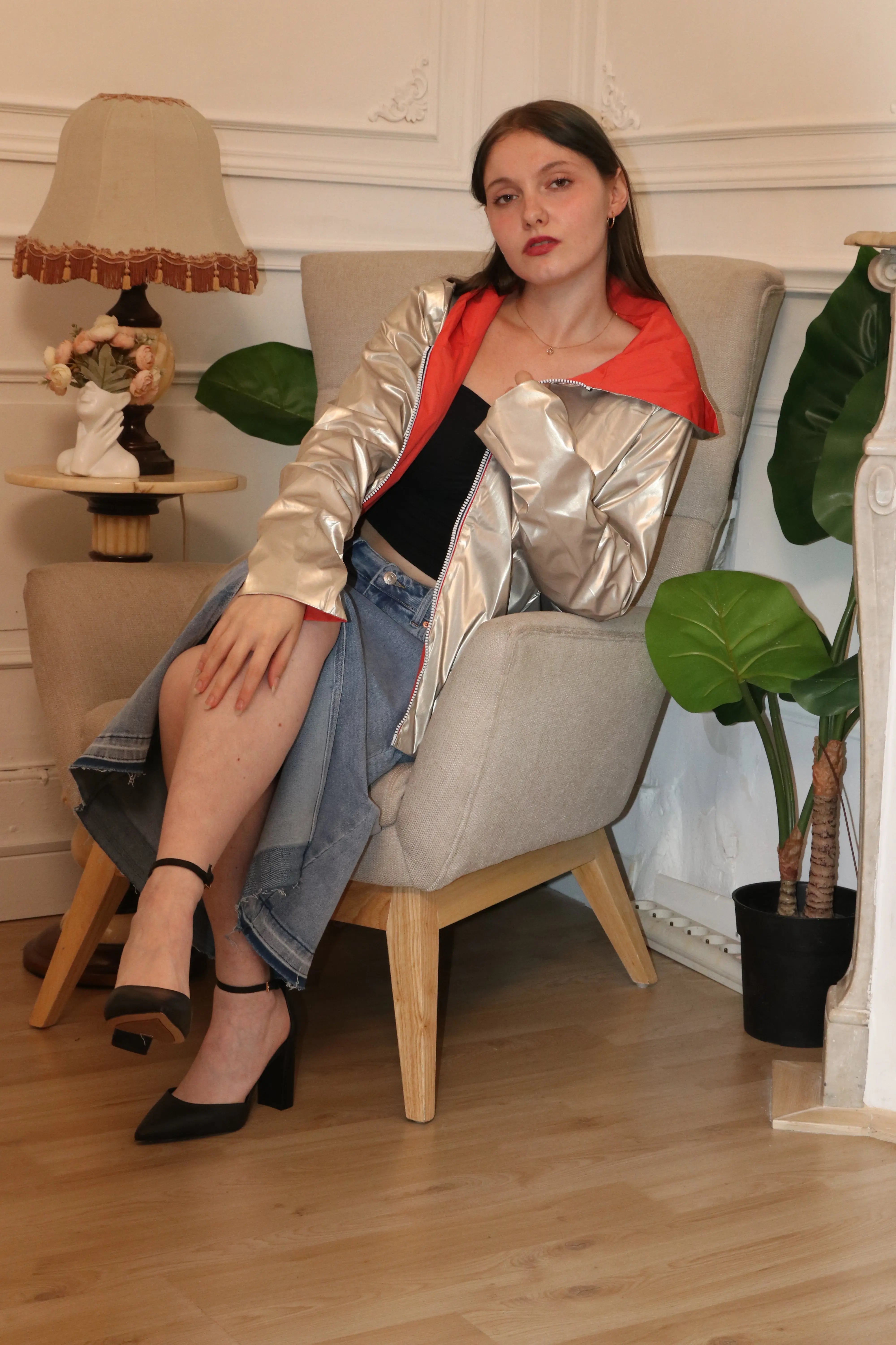 Femme portant une veste argentée métallisée et une jupe en jean assise dans un fauteuil.