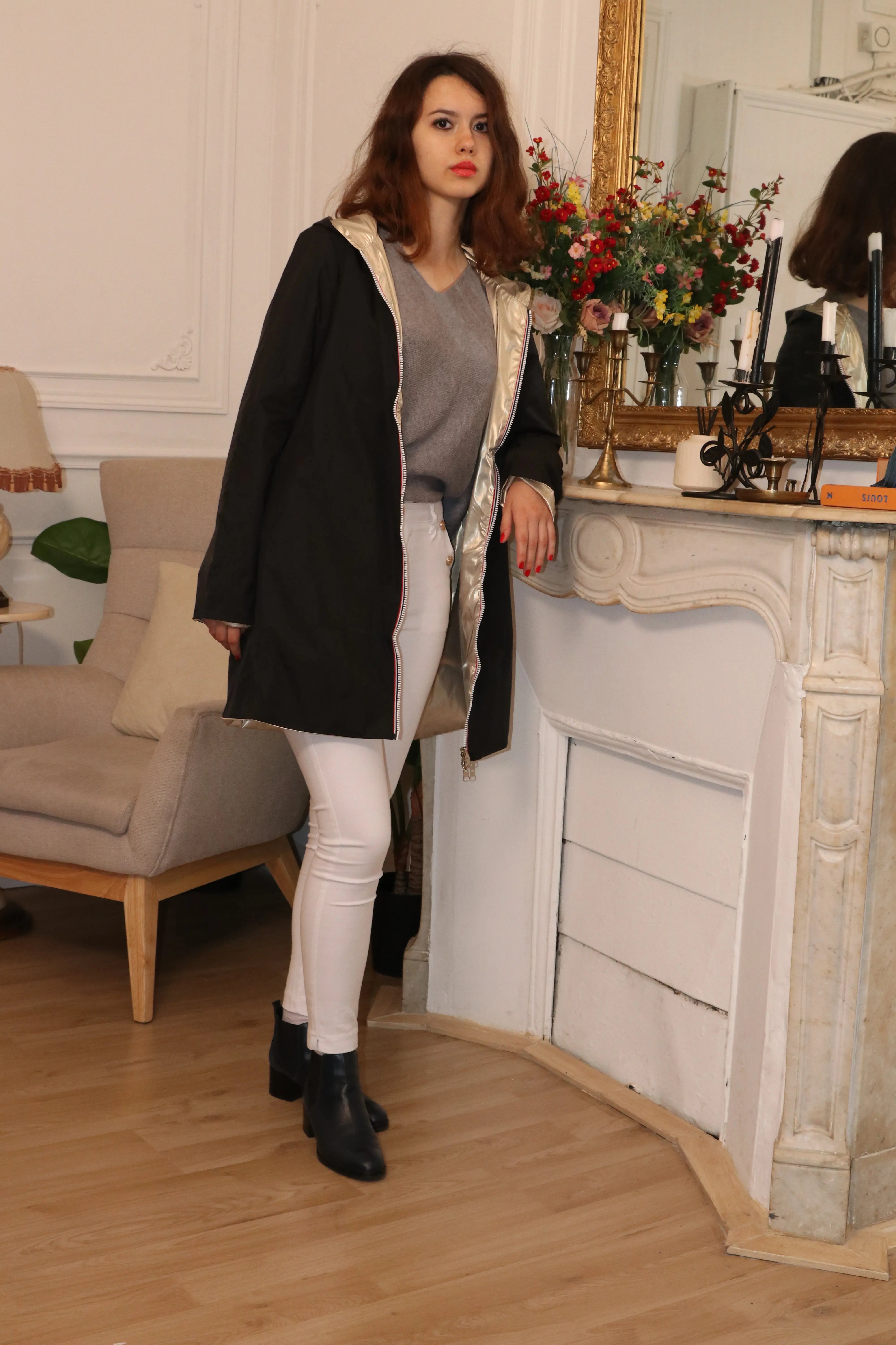Femme portant un manteau noir élégant, un pull gris et un pantalon blanc près d'une cheminée.