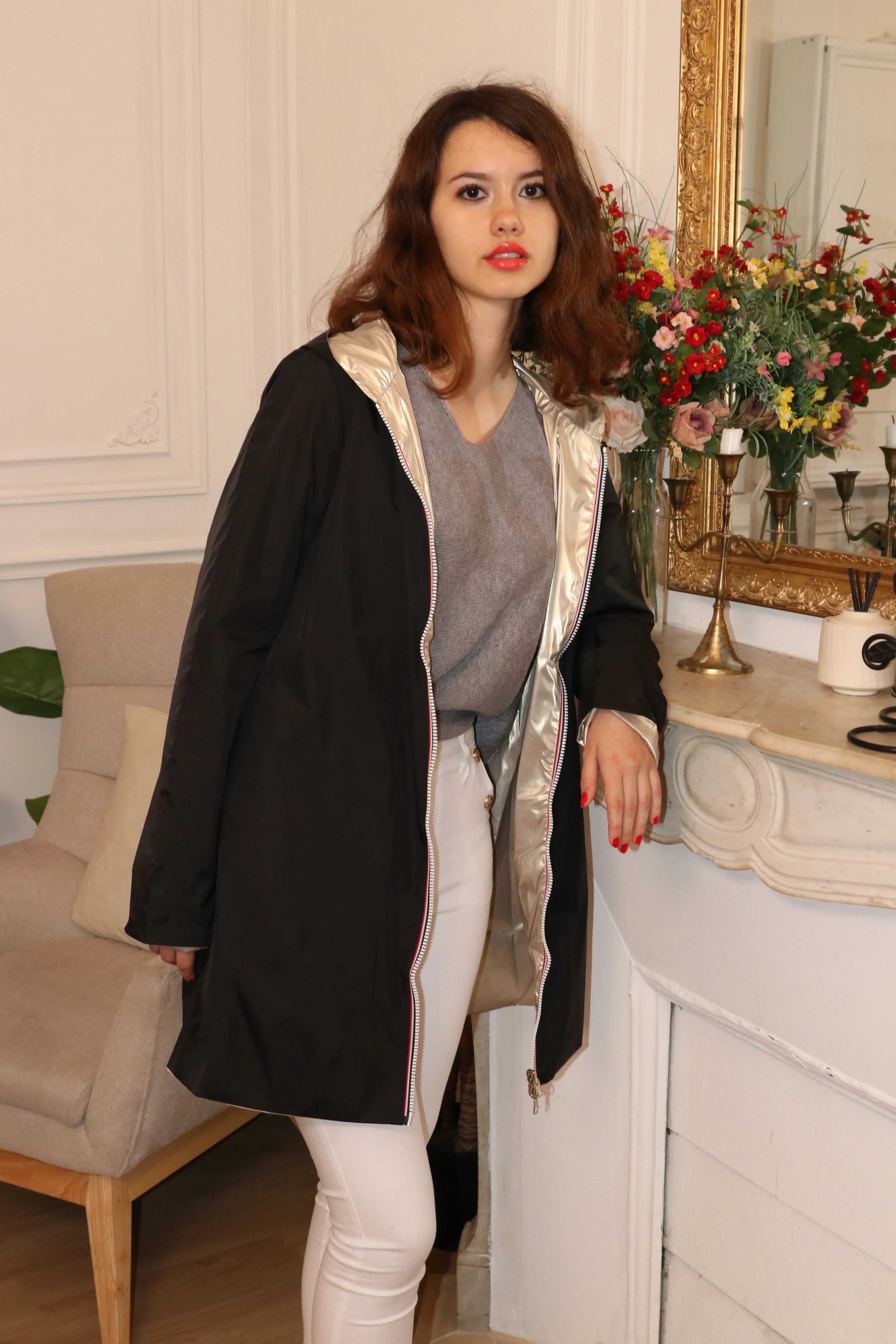 Femme portant un manteau noir ouvert sur un pull gris et un pantalon blanc.