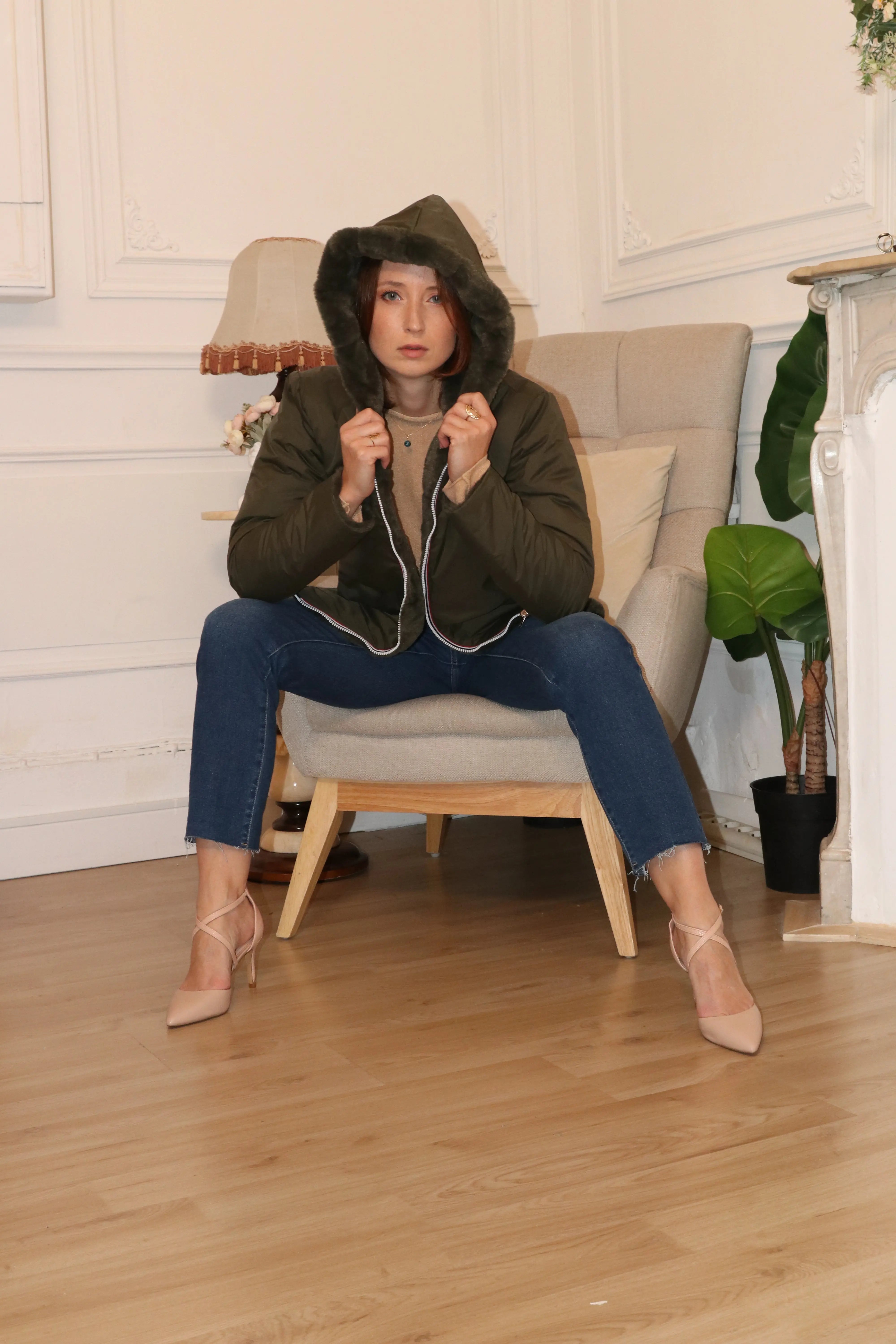 Femme portant une doudoune kaki à capuche et un jean bleu assise dans un fauteuil.