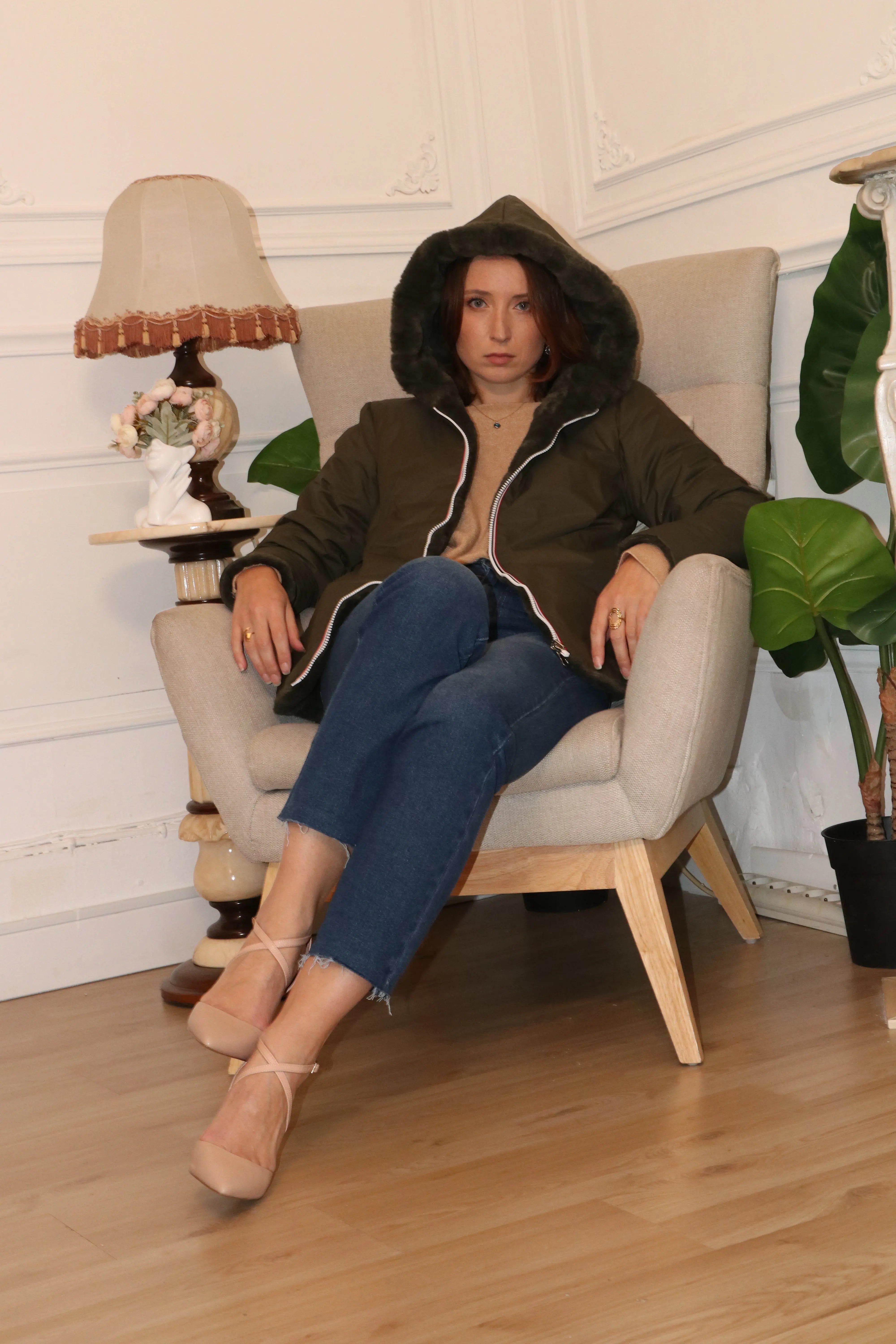 Femme portant une parka à capuche olive et un jean bleu assise dans un fauteuil beige.
