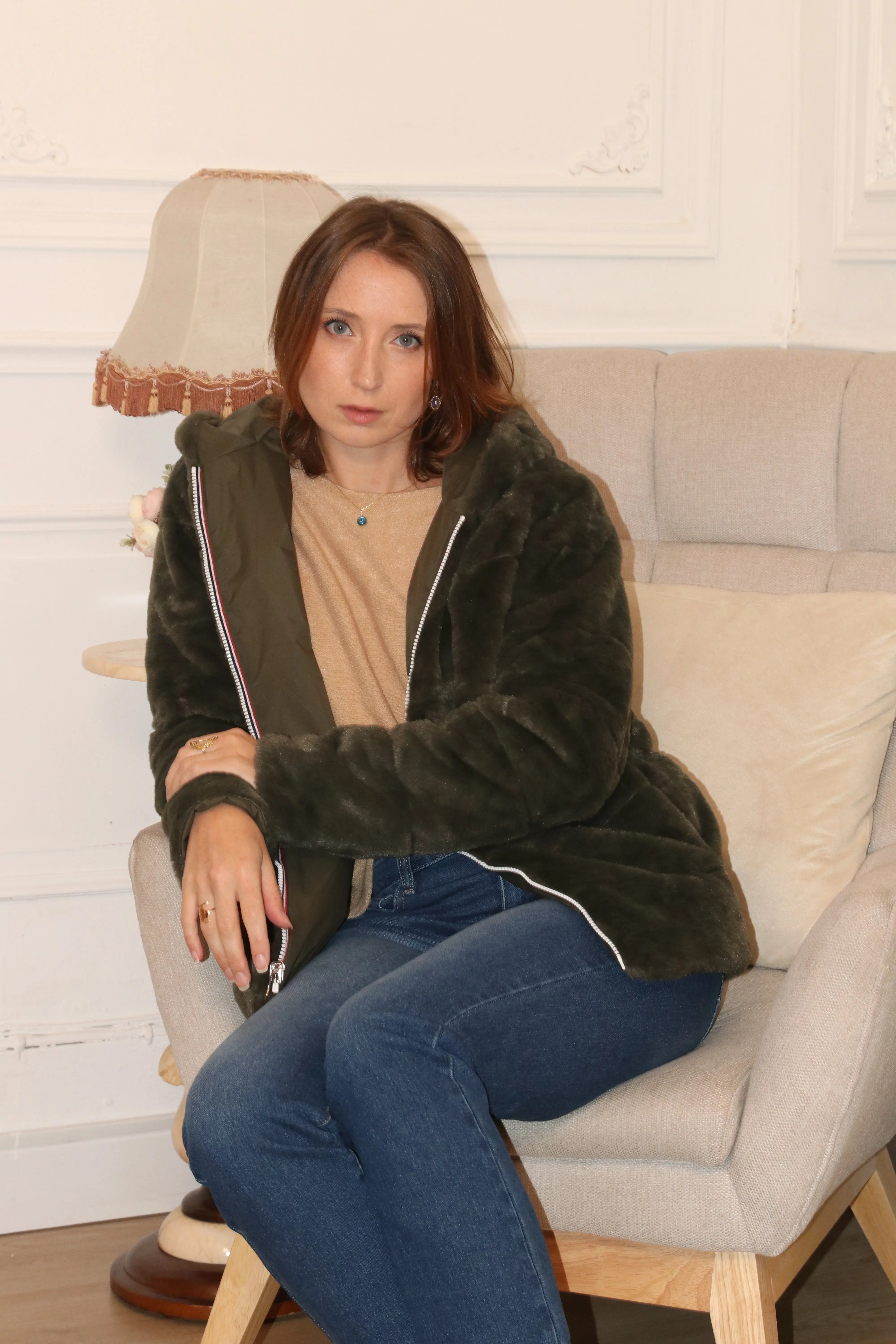 Femme portant une veste en fausse fourrure vert olive et un jean bleu assise dans un fauteuil.