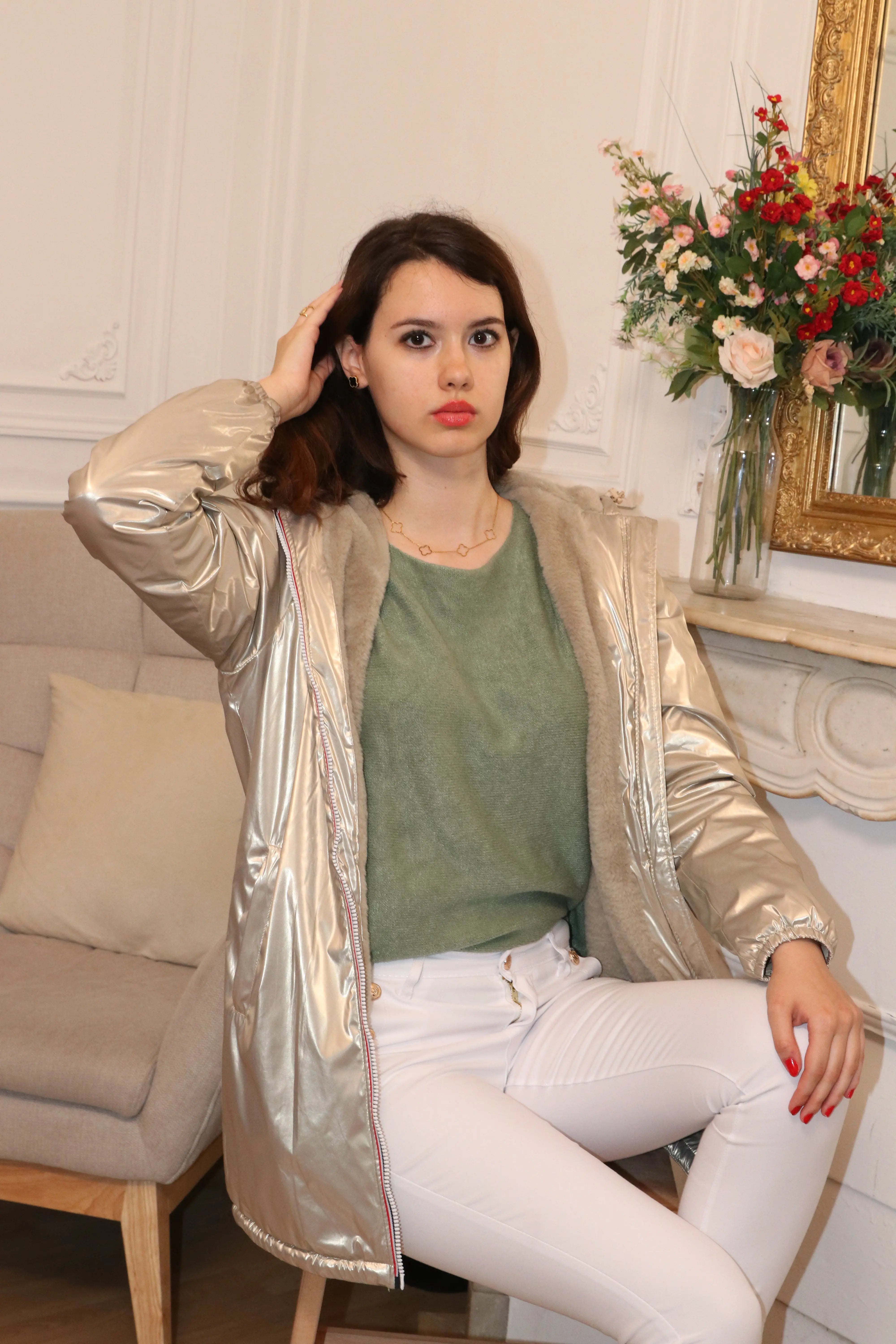 Femme portant une veste métallisée argentée et un pull vert olive sur un canapé gris.