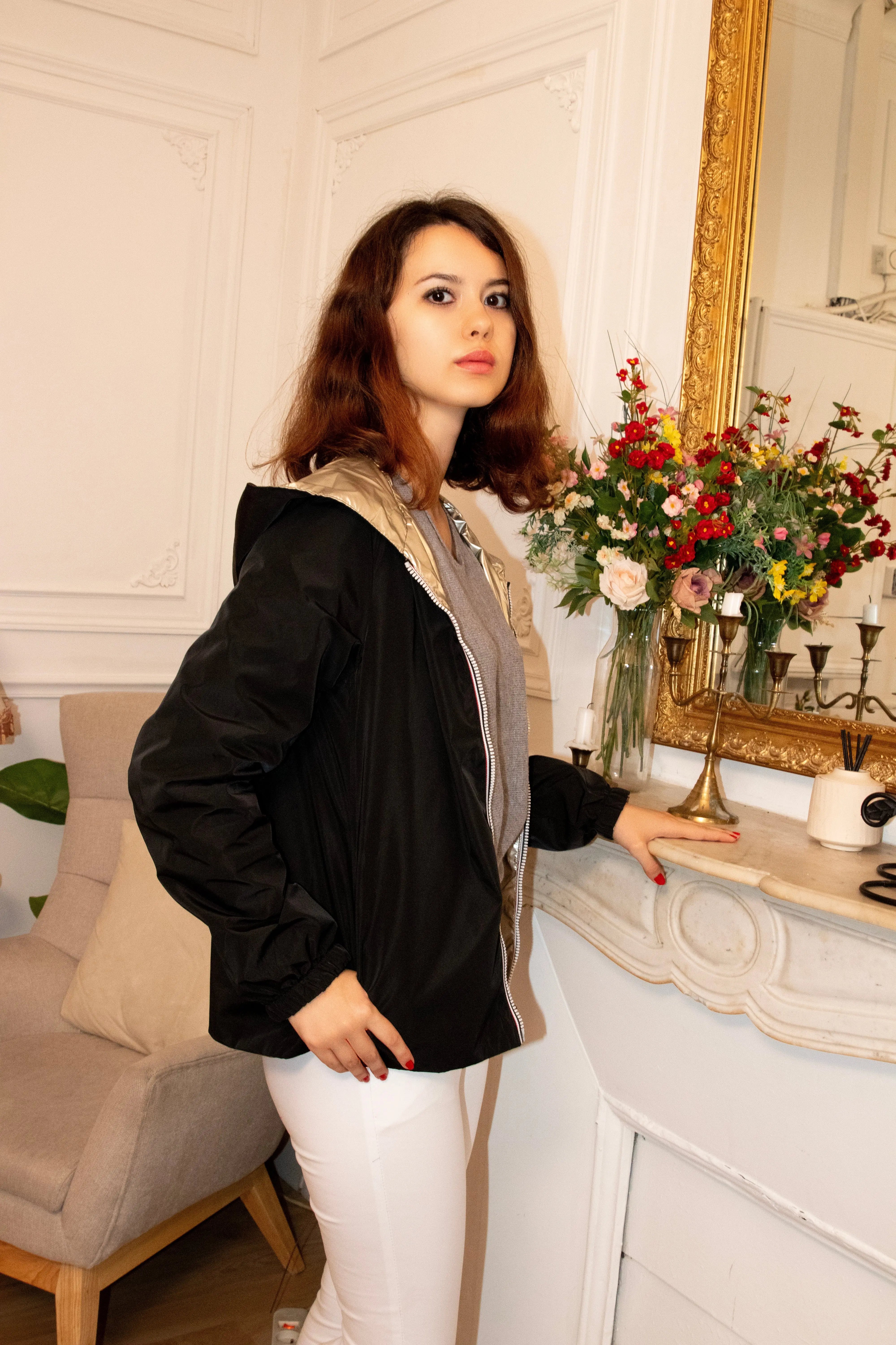 Femme portant une veste noire et dorée et un pantalon blanc devant une cheminée fleurie.