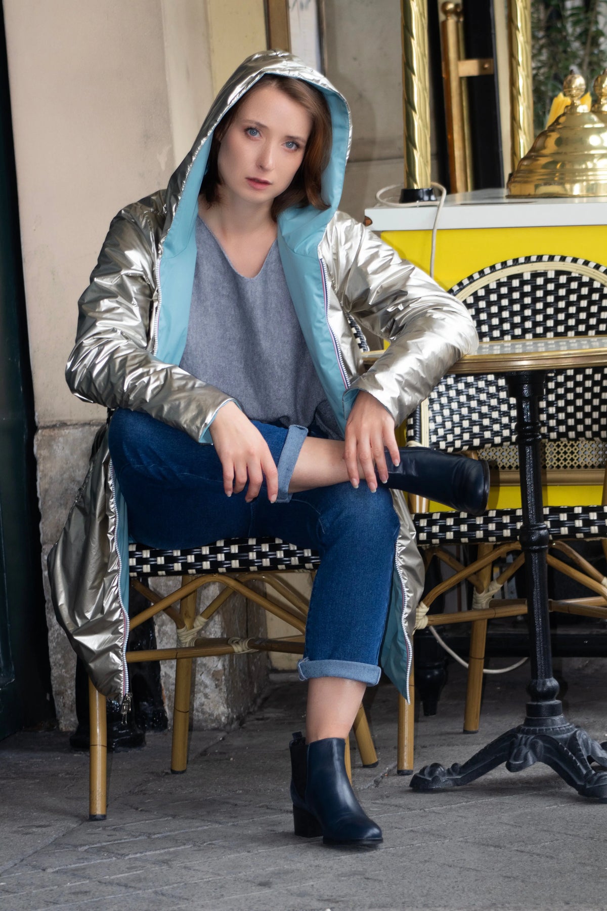 Femme portant un manteau argenté brillant avec capuche et jean bleu en terrasse.